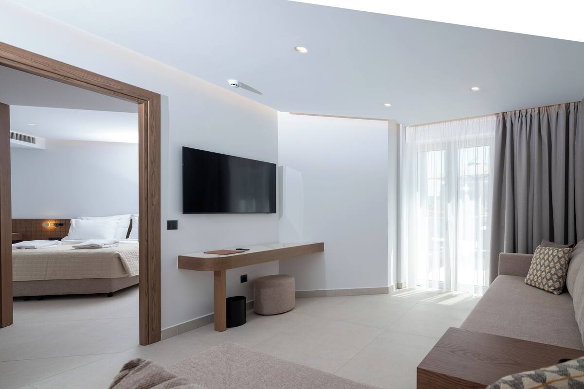 A modern hotel room with a bed, a sofa, a TV mounted on the wall, and a large window with curtains.