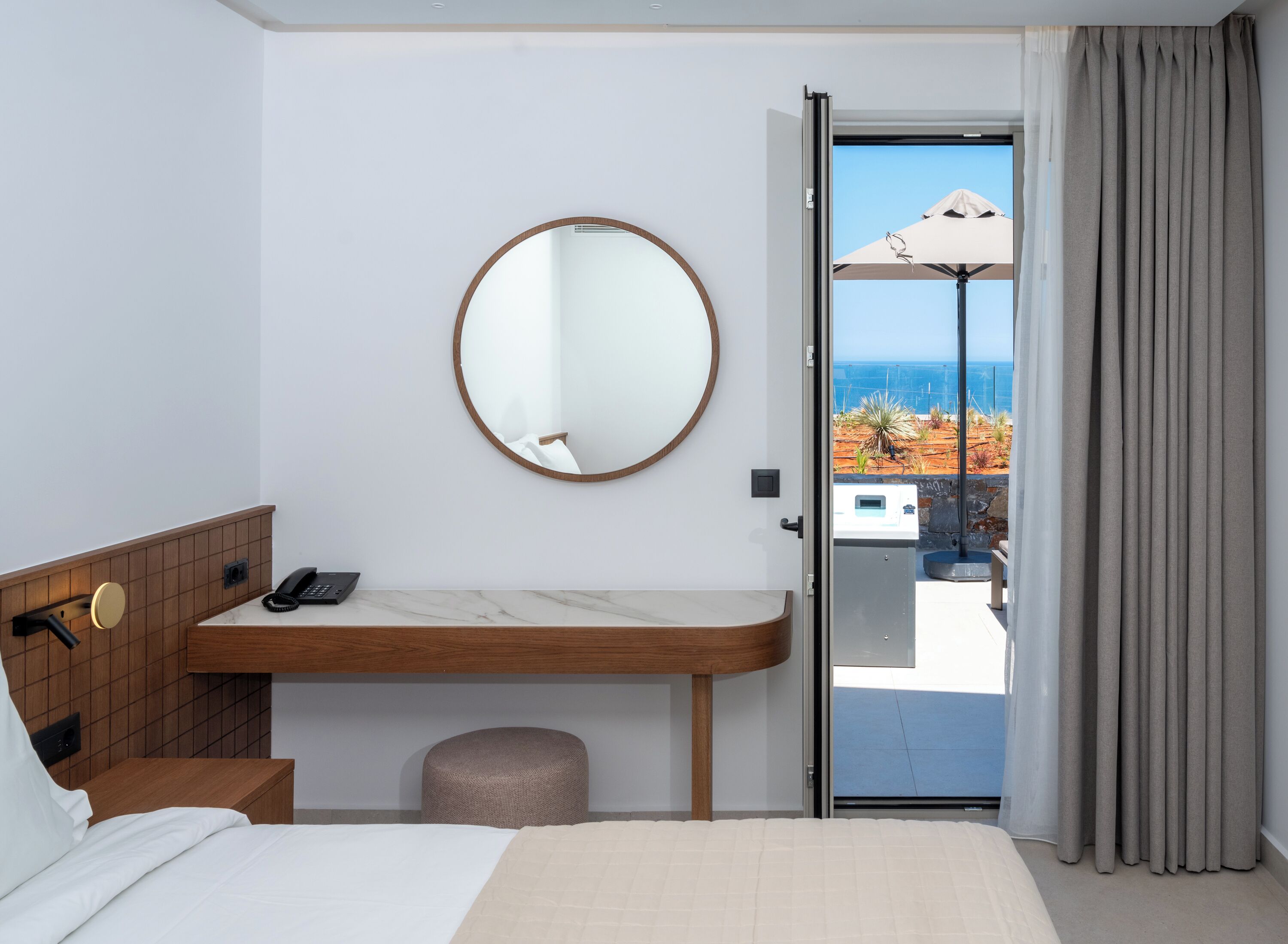 Une chambre d’hôtel avec un lit, une table de chevet en bois avec un téléphone, un miroir rond et une vue sur la mer par une porte vitrée.