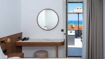 Une chambre d’hôtel avec un lit, une table de chevet en bois avec un téléphone, un miroir rond et une vue sur la mer par une porte vitrée.
