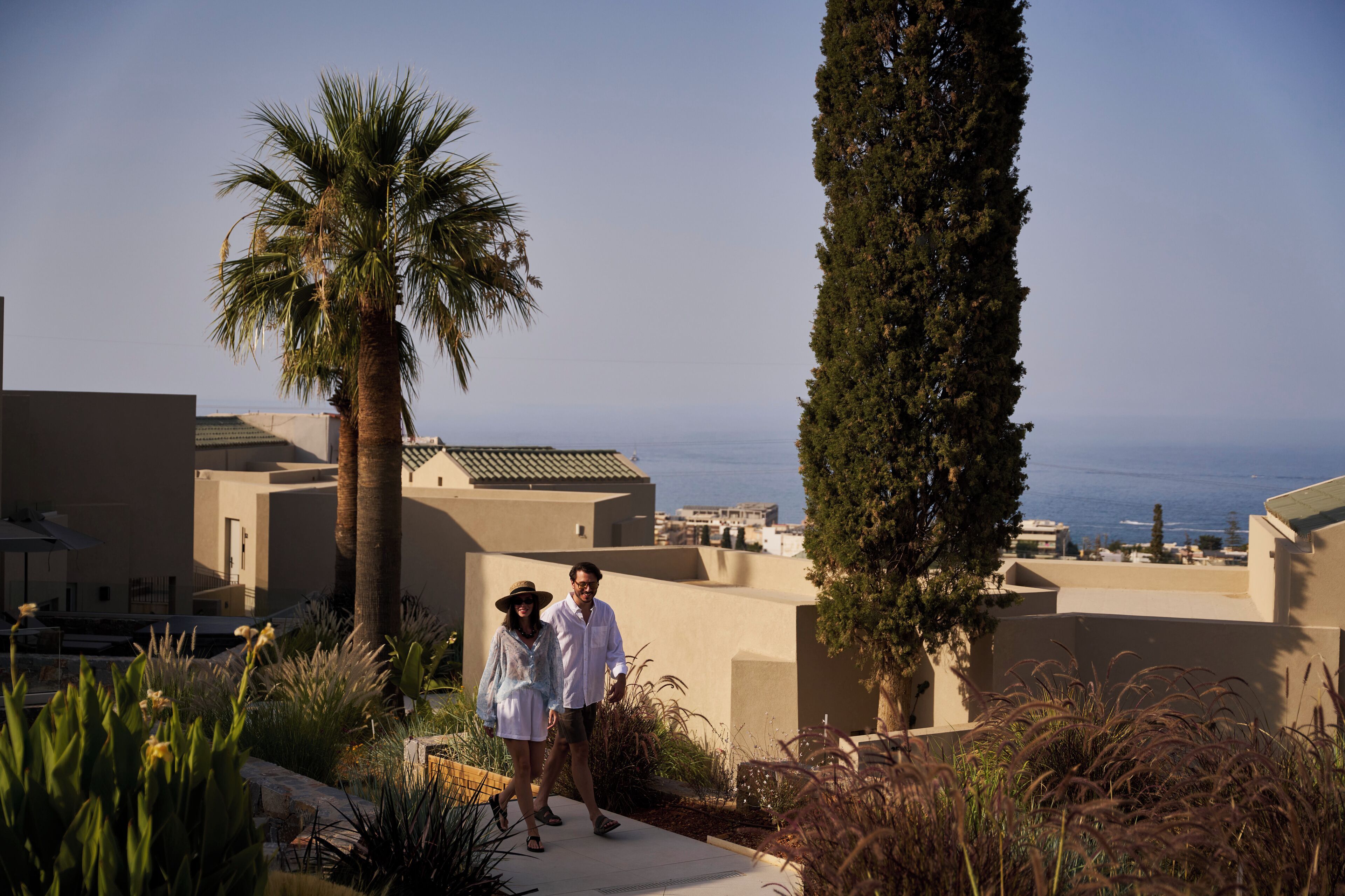 A couple walking on a pathway with a view of the sea and buildings in the background.