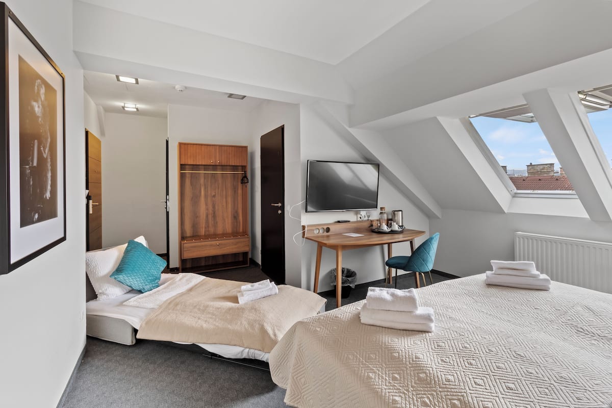 A modern hotel room with a loft-style ceiling, a bed with a patterned duvet, a desk with a flat-screen TV, and a view of the city through a skylight.