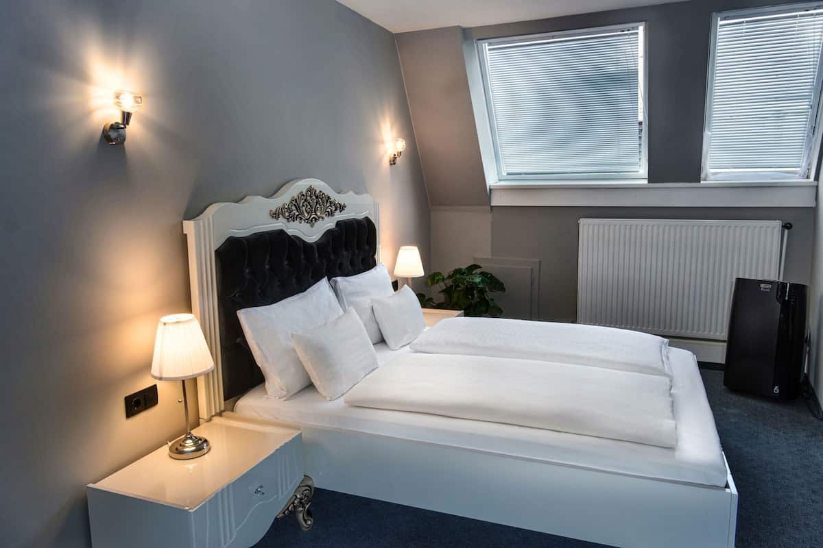 Un lit bien fait, avec du linge de lit blanc, deux lampes de chevet, un radiateur et une fenĂȘtre avec des stores.