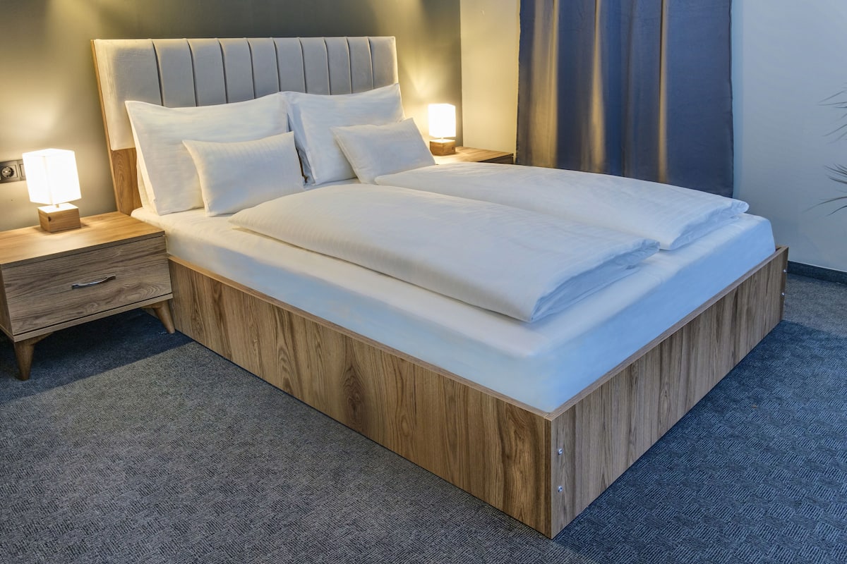 A neatly made bed with a wooden frame and a headboard, accompanied by two bedside tables with lamps, in a room with a carpeted floor.