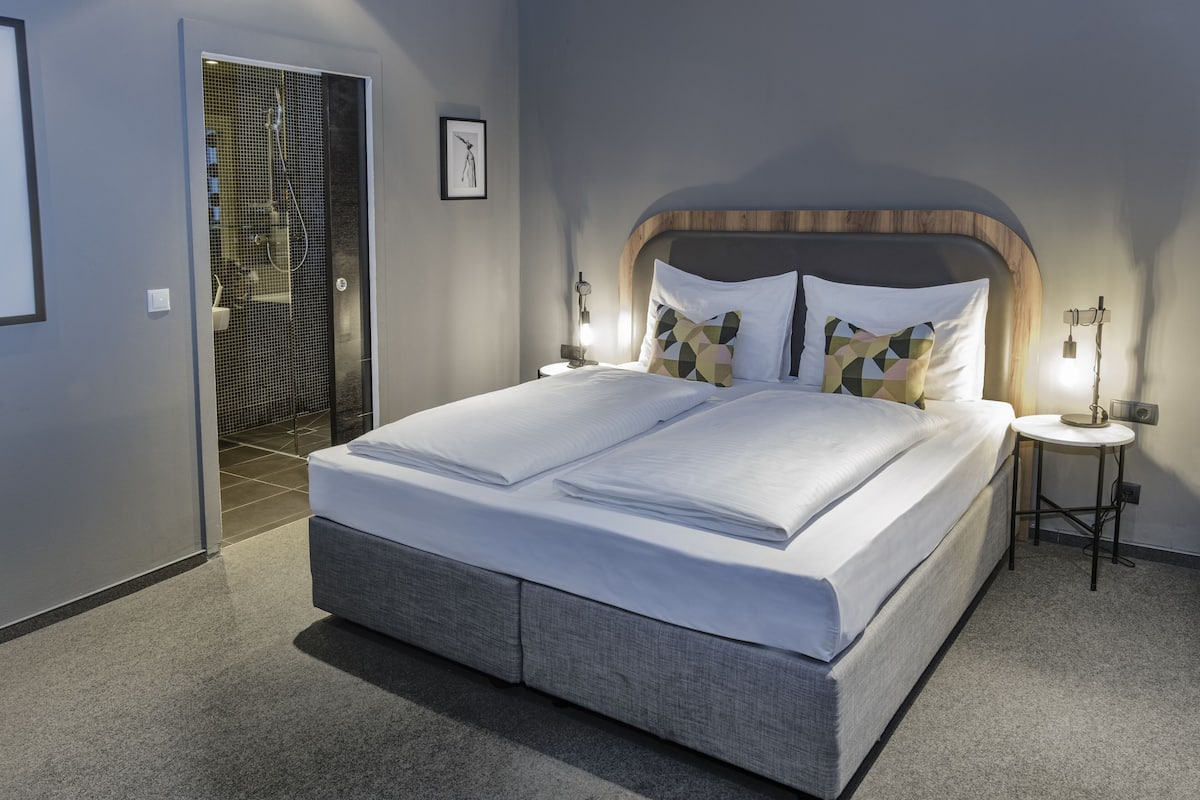 A modern bedroom with a large bed, a bedside table with a lamp, and a shower area visible through a glass door.