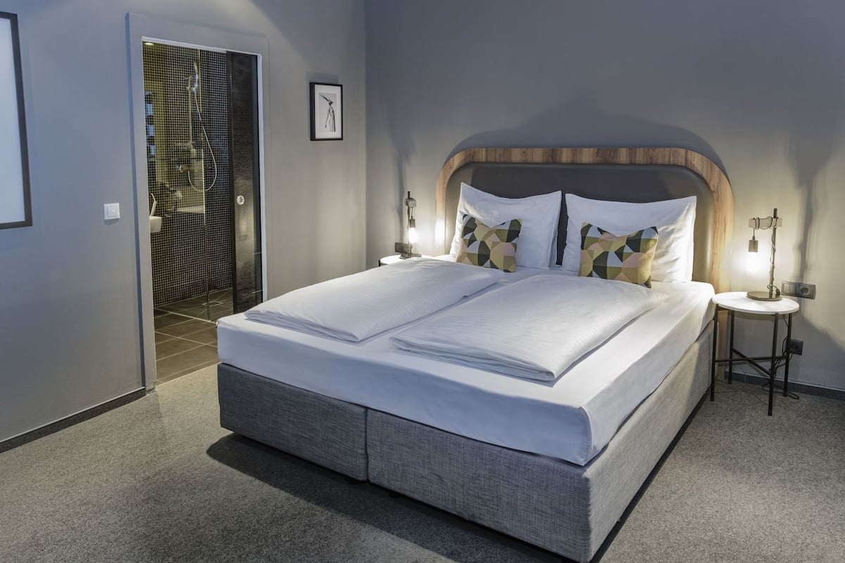 A modern bedroom with a large bed, a bedside table, a wall-mounted light, and a shower area.
