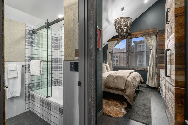 A modern bathroom with a glass-enclosed shower and a separate bedroom with a large bed and a view of the city.