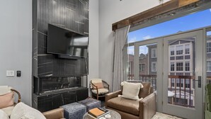 Ein modernes Wohnzimmer mit großem Flachbildfernseher, bequemen Sitzmöglichkeiten, einem Kamin und Blick auf die Stadt durch bodentiefe Fenster.