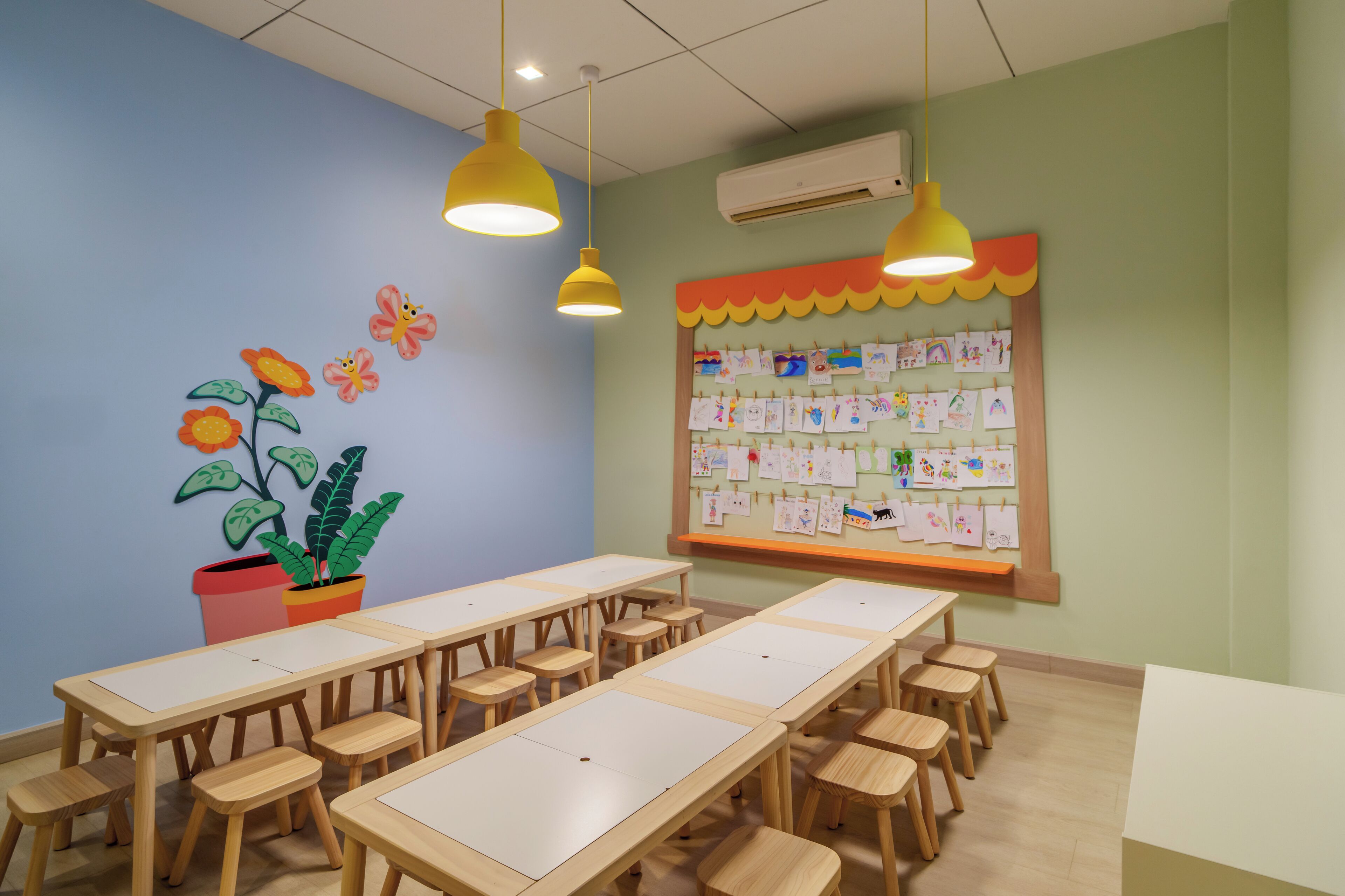 A classroom with wooden tables and chairs, a bulletin board with children's drawings, and a potted plant.
