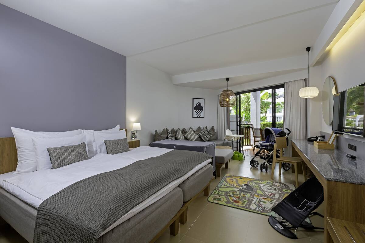 Una habitación de hotel moderna con una cama grande, un escritorio, una silla, un cochecito de bebé y vistas al exterior.