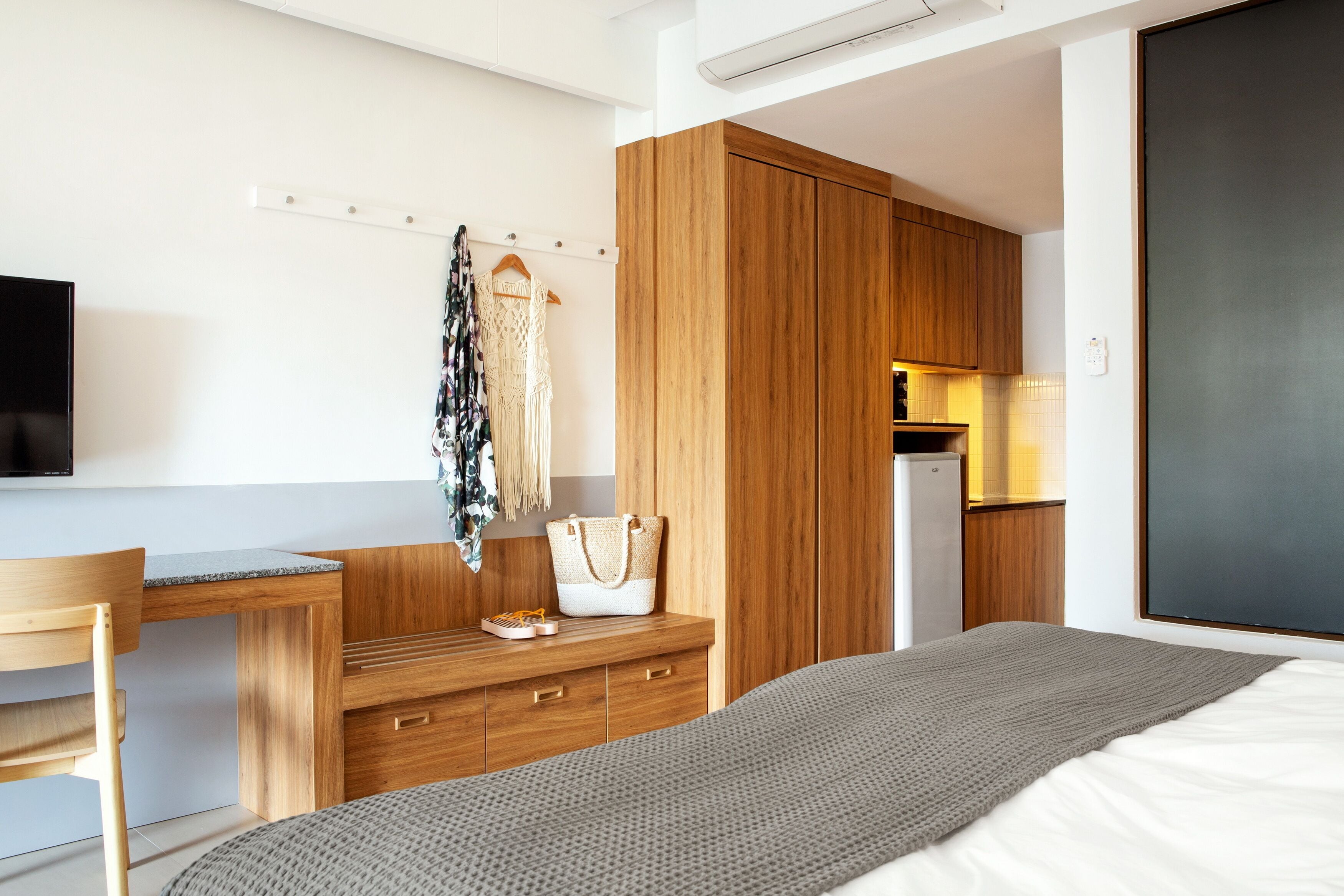 A modern hotel room with a wooden desk, a bed, a wardrobe, and a kitchenette.