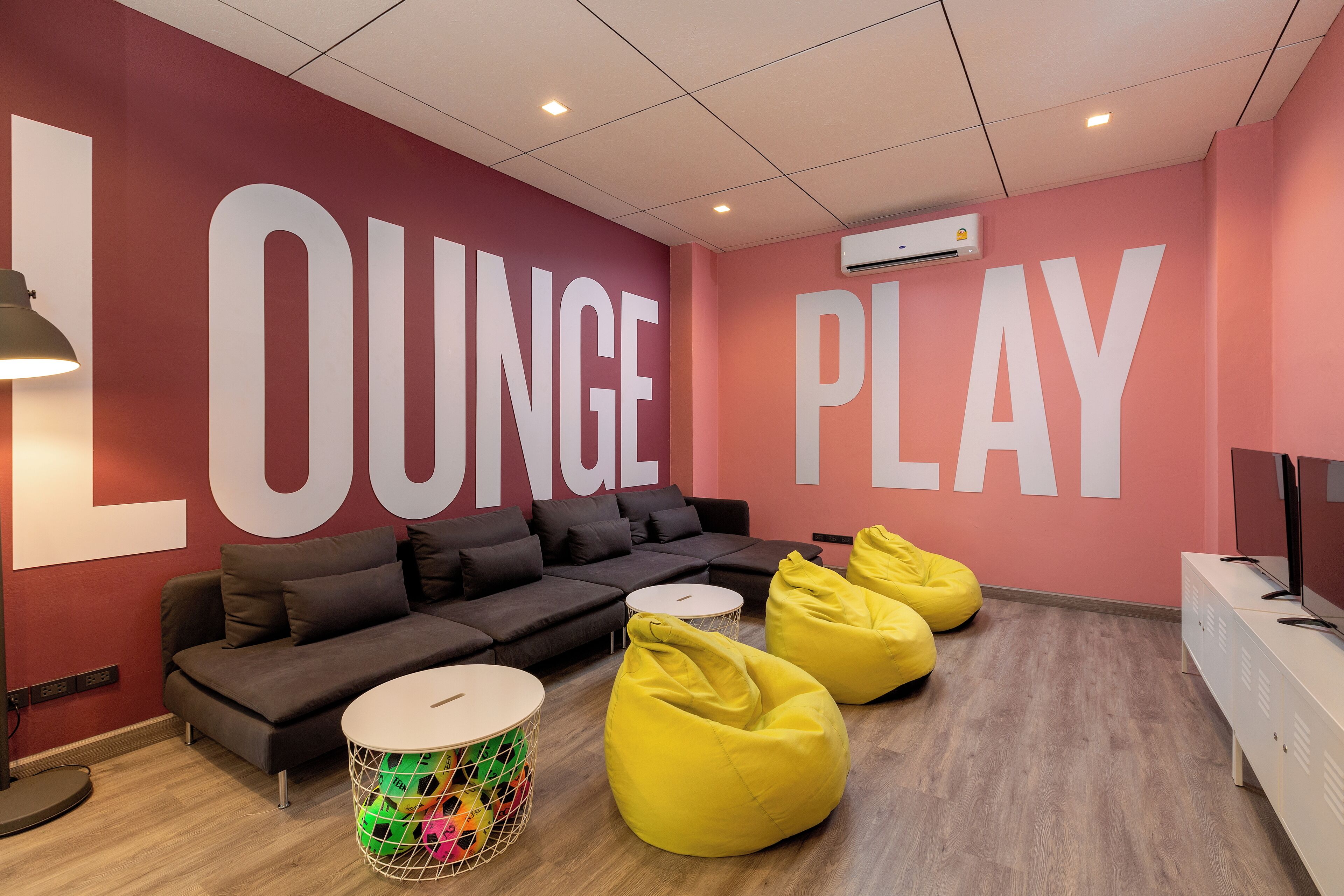 Un área de descanso con un letrero grande que dice 'LOUNGE PLAY', un sofá, sillones de mimbre amarillos, una pequeña mesa redonda y una cesta colorida.