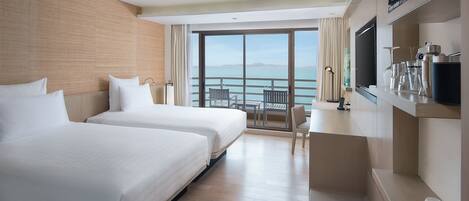 A hotel room with two beds, a desk, a TV, and a balcony with a view of the sea.