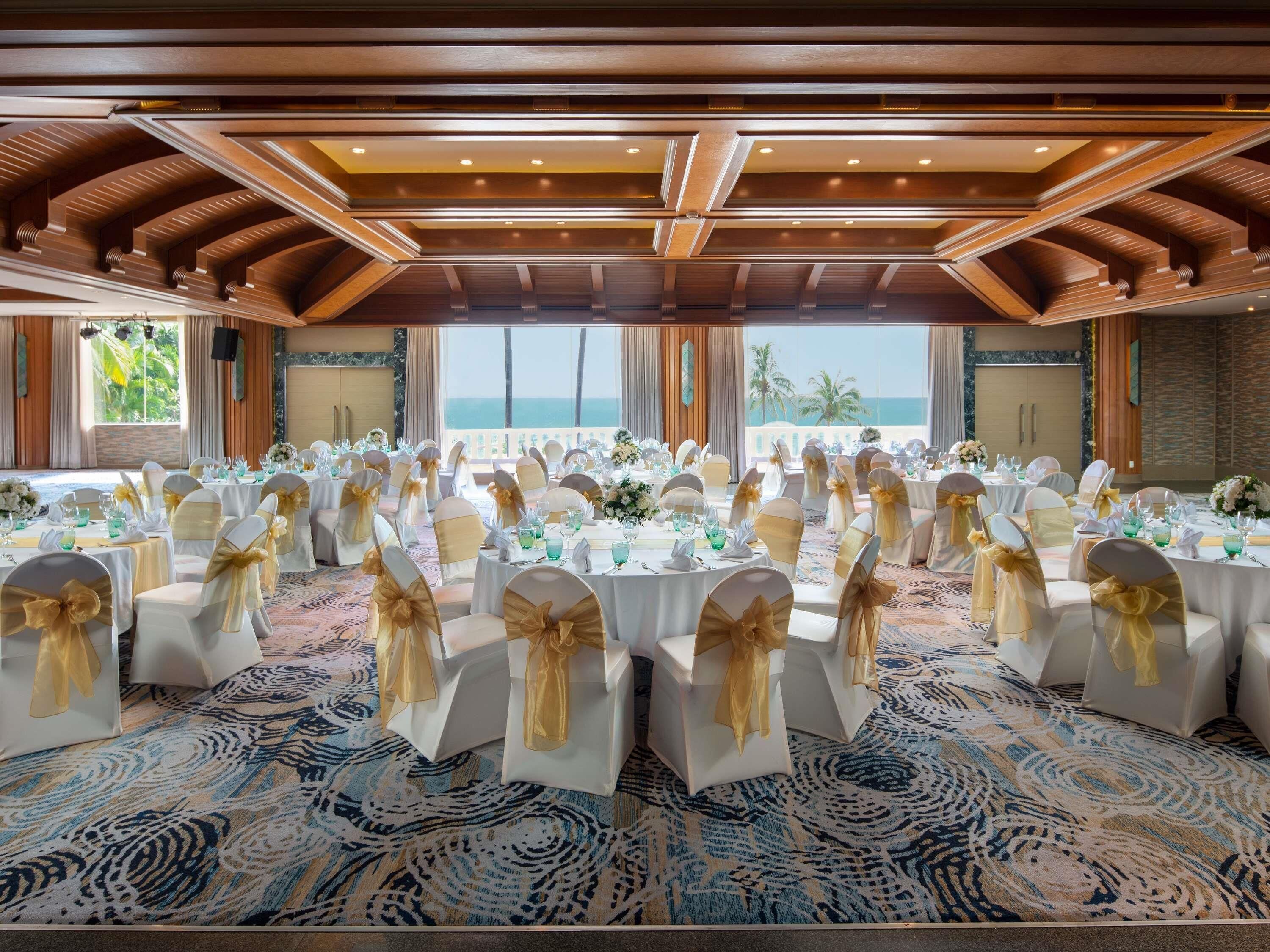 Een feestzaal met ronde tafels, witte stoelen met gouden linten en uitzicht op de oceaan.
