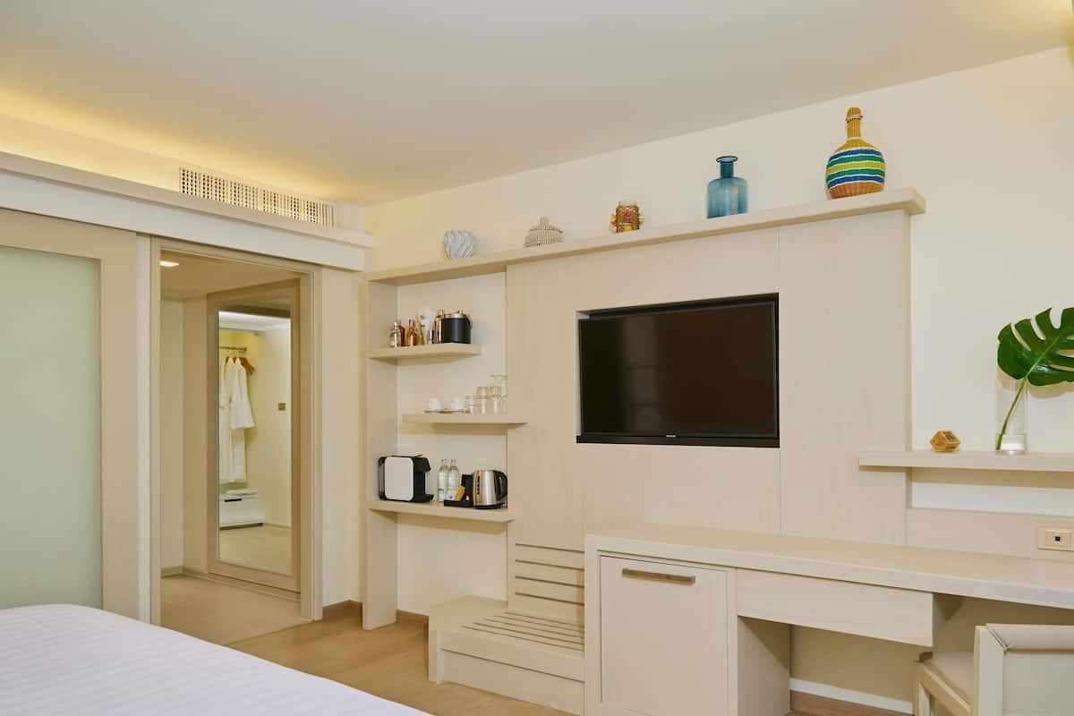 A hotel room with a bed, a desk with a chair, a TV, and a shelf with various items.