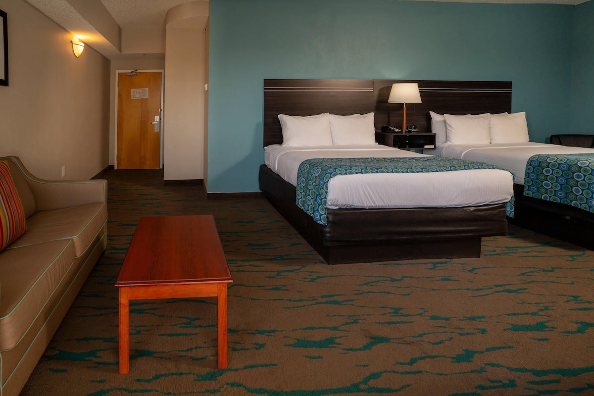 A hotel room with two beds, a wooden nightstand, a lamp, a brown leather sofa, and a patterned carpet.