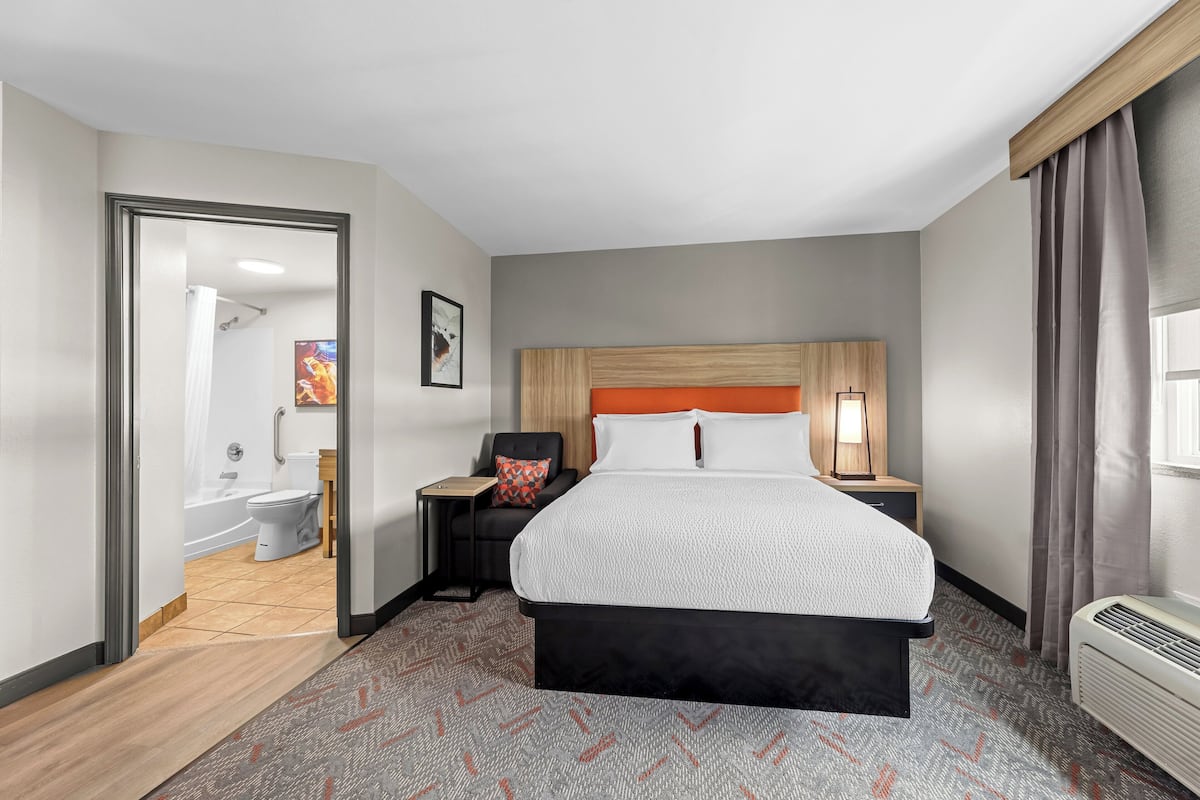 A hotel room with a large bed, a nightstand, a chair, a bathroom with a bathtub, and a window with curtains.