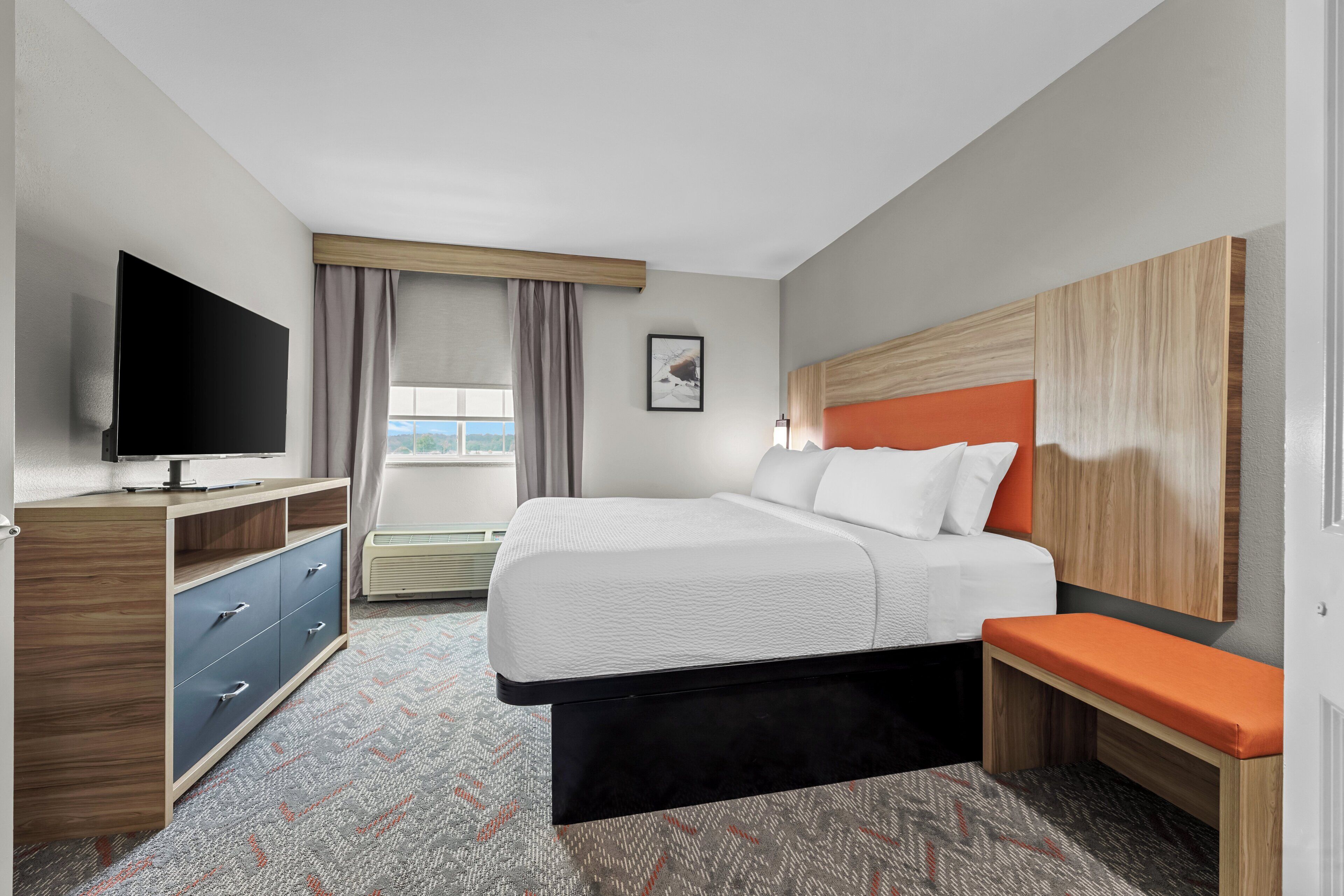 A modern hotel room with a bed, a TV, a desk, and a window with curtains.