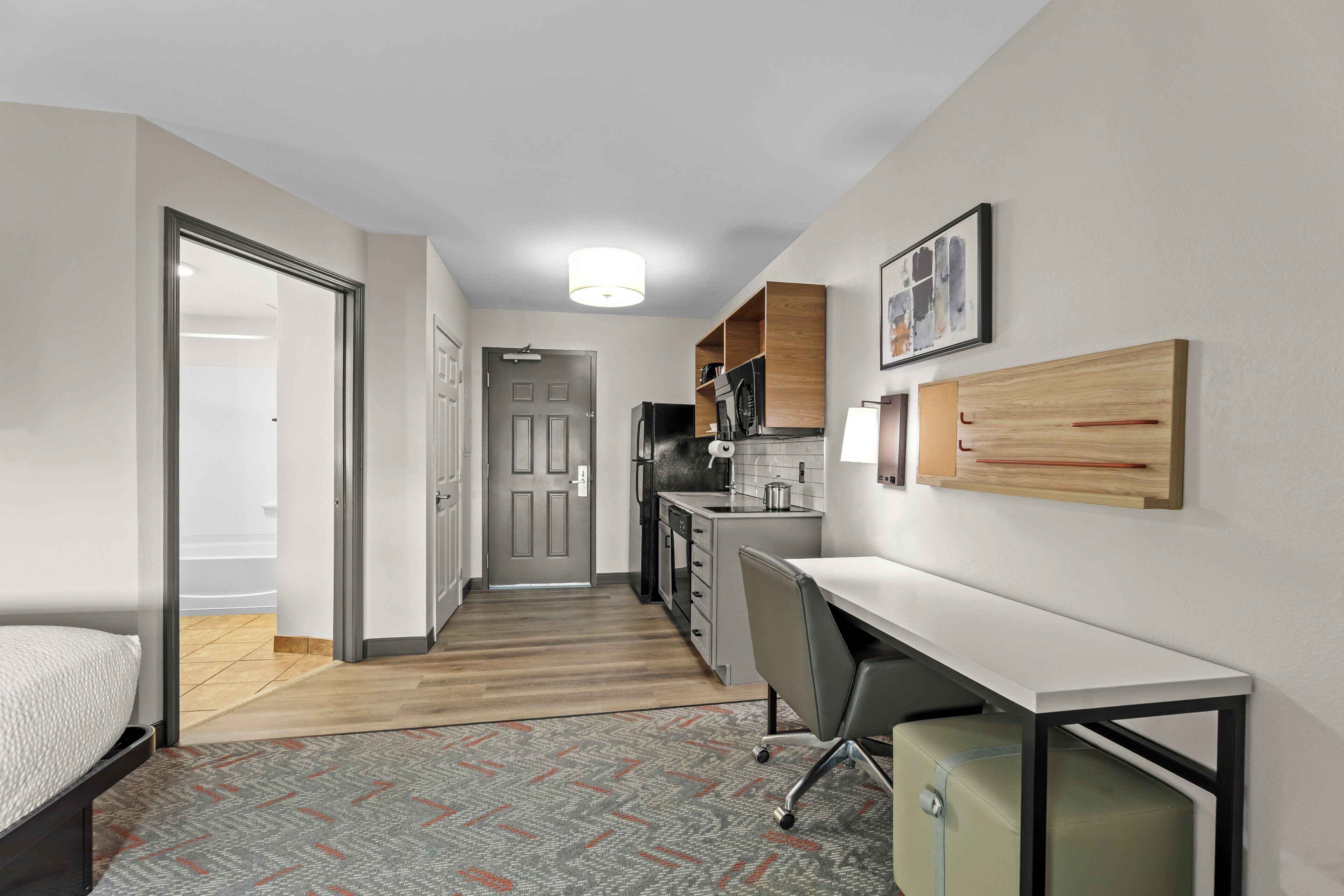 A compact hotel room with a bed, a desk, a chair, a kitchenette, and a bathroom.