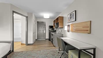 A compact hotel room with a bed, a desk, a chair, a kitchenette, and a bathroom.