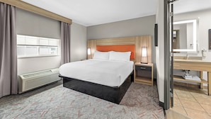 Una habitación de hotel con cama, mesitas de noche, tocador, espejo, baño con lavamanos e inodoro, y ventana con cortinas.