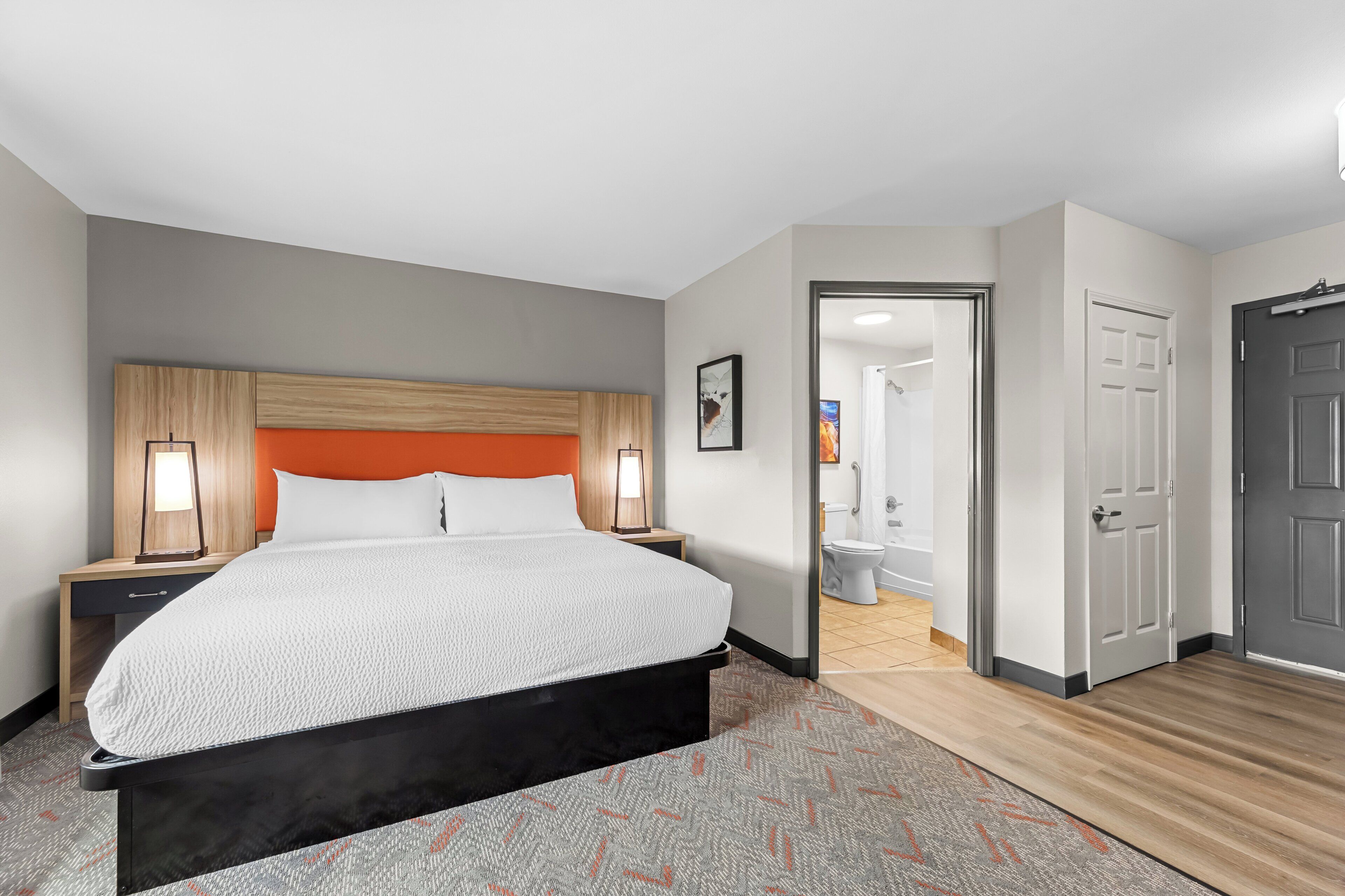Ein Hotelzimmer mit einem Bett, Nachttischlampen, einem Kopfteil, einer Tür und einem durch eine geöffnete Tür einsehbaren Badezimmer.