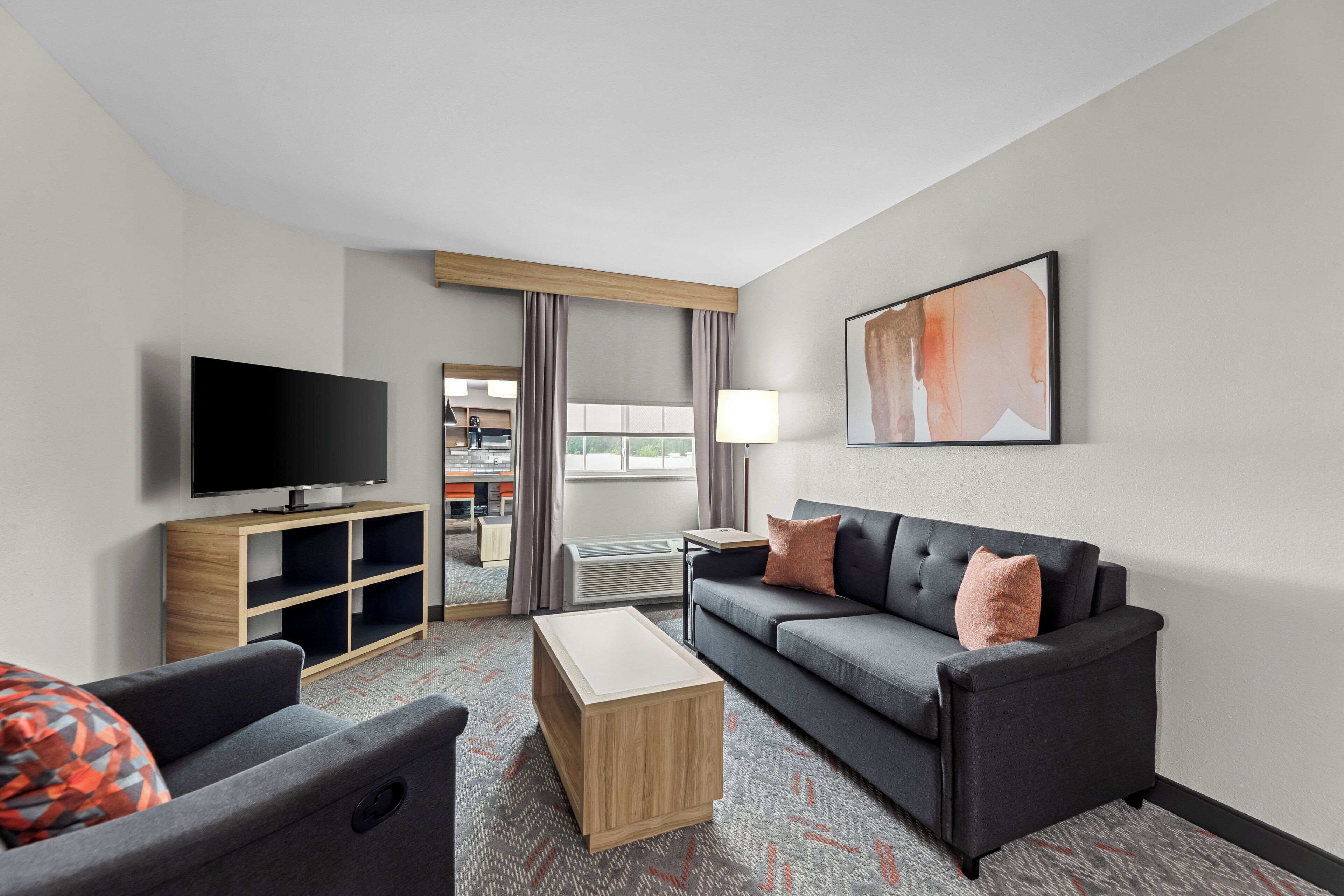 A modern hotel room with a flat-screen TV, a sofa, a coffee table, and a view of the kitchen area.