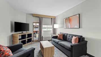 A modern hotel room with a flat-screen TV, a sofa, a coffee table, and a view of the kitchen area.