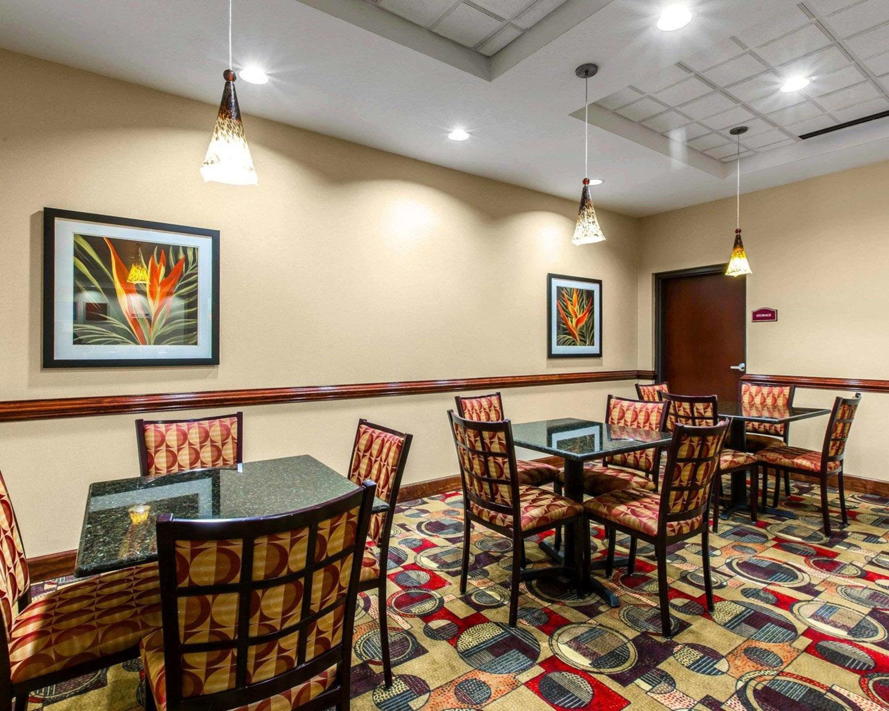 A hotel room with a dining area featuring a patterned carpet, multiple tables and chairs, and two wall-mounted pendant lights.