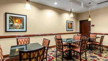 A hotel room with a dining area featuring a patterned carpet, multiple tables and chairs, and two wall-mounted pendant lights.