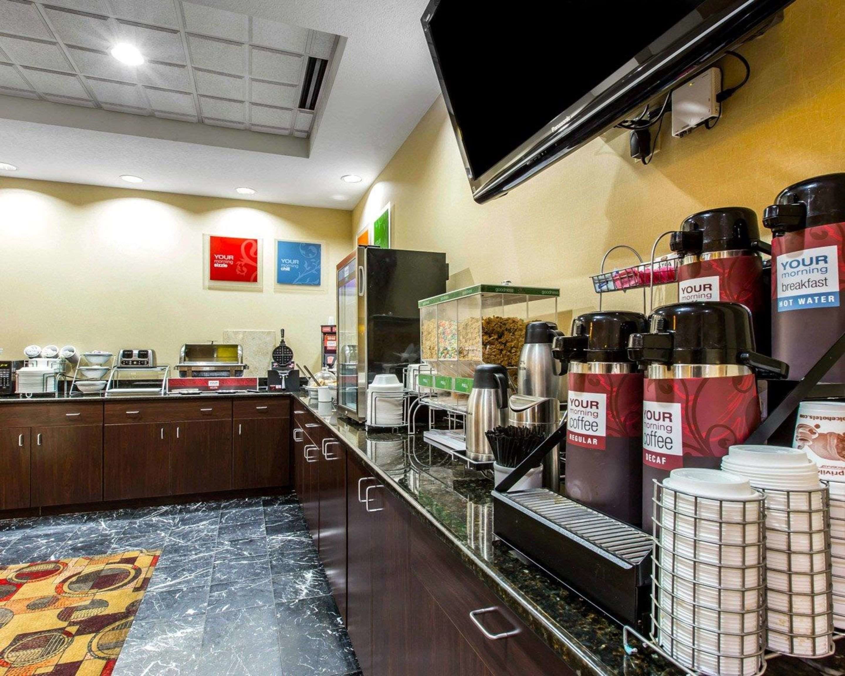 A hotel breakfast area with a self-service coffee station, a refrigerator, and a flat-screen TV mounted on the wall.
