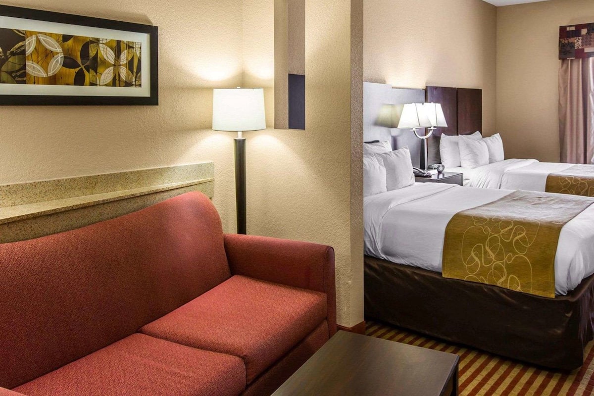 Quarto de hotel com sofá vermelho, cama com roupa de cama branca, mesa de cabeceira com abajur e um quadro na parede.