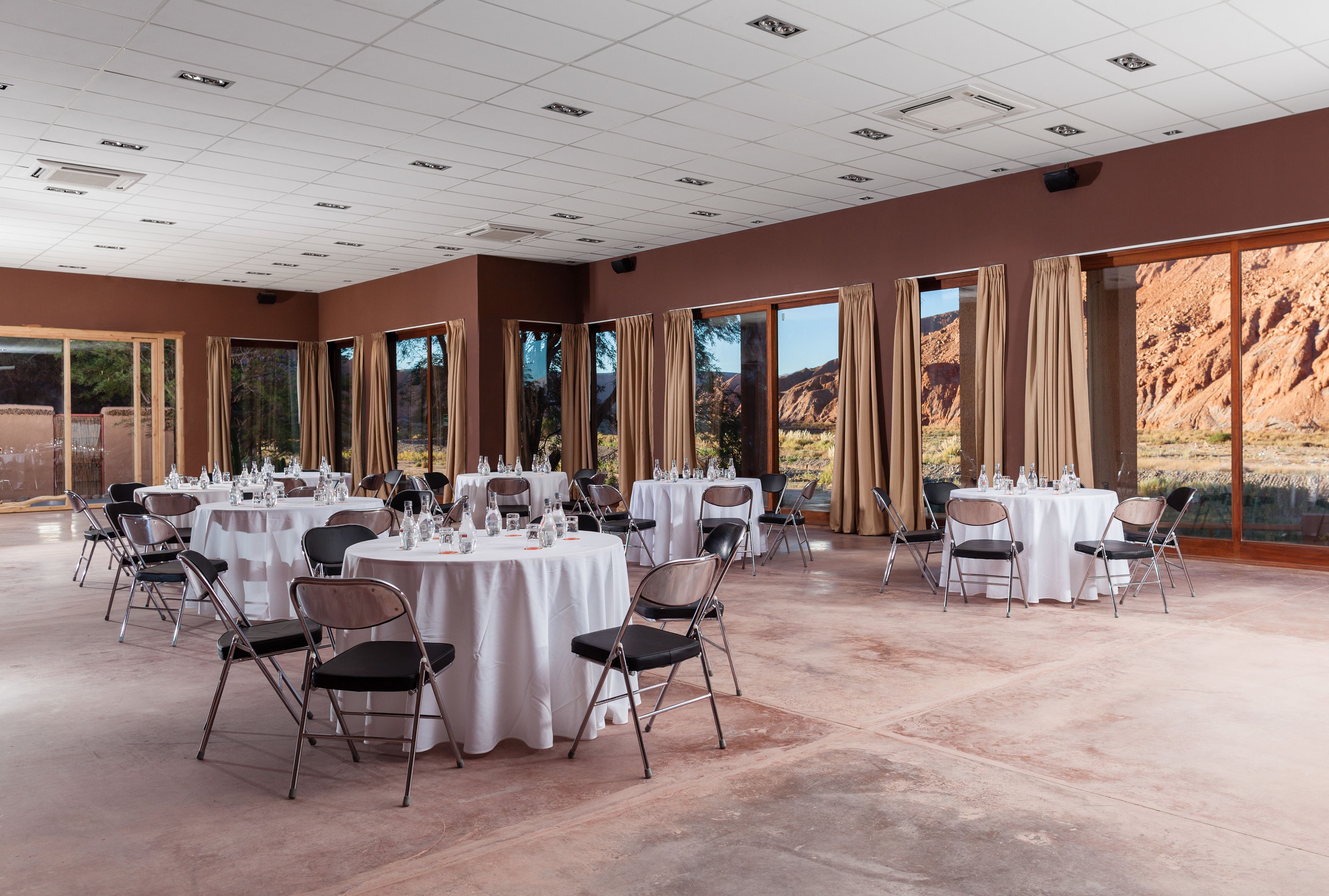 Un vaste espace intérieur doté de plusieurs tables et chaises rondes, de grandes fenêtres avec des rideaux et offrant une vue sur un paysage montagneux.