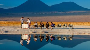 Un grupo de personas sentadas en sillas junto a una superficie acuática reflectante, con montañas al fondo.