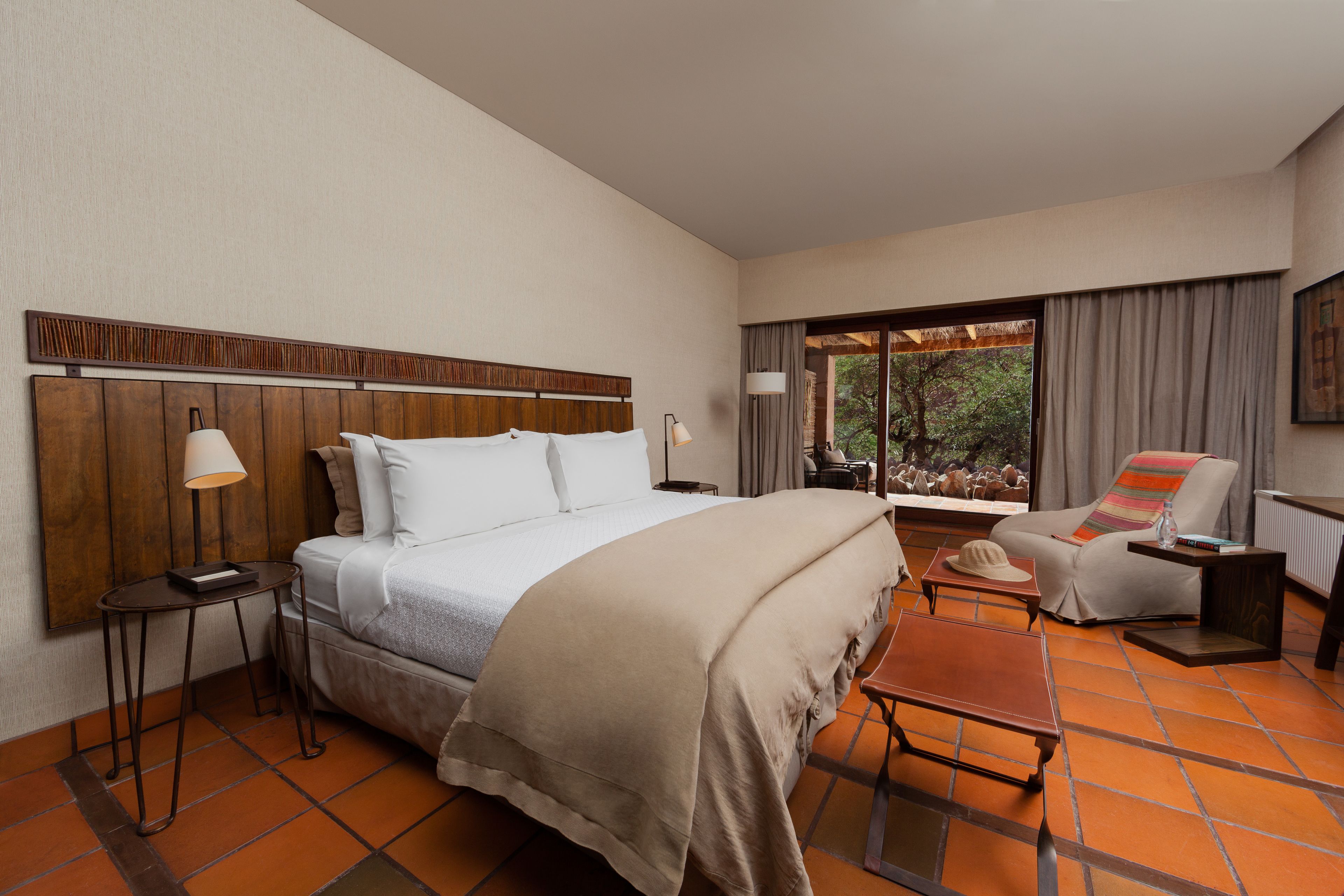 Una habitación de hotel con una cama grande, un área de descanso con una silla y una mesa, una mesita auxiliar pequeña y un ventanal con cortinas.