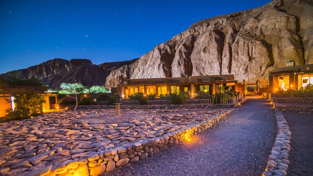 Un hotel con un camino de piedra que lleva a un edificio con múltiples ventanas, con montañas al fondo.