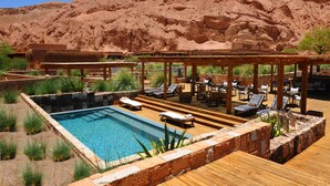 Un área de piscina con sillones de descanso y una terraza de madera, rodeada de vegetación desértica y con montañas al fondo.