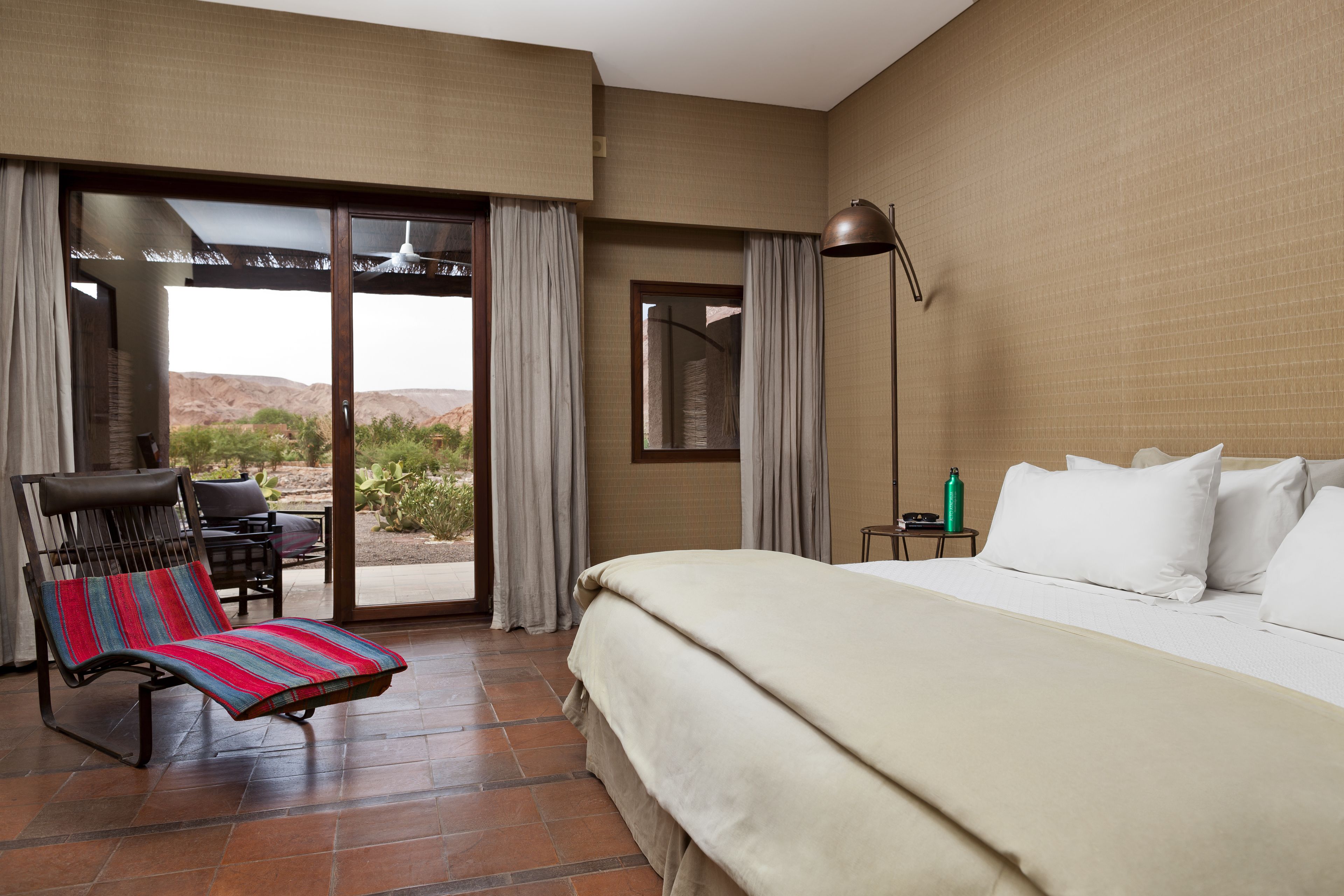 Une chambre d’hôtel avec un lit, une chaise dotée d’un coussin à rayures, une table et une vue sur le désert, visible à travers une porte coulissante.