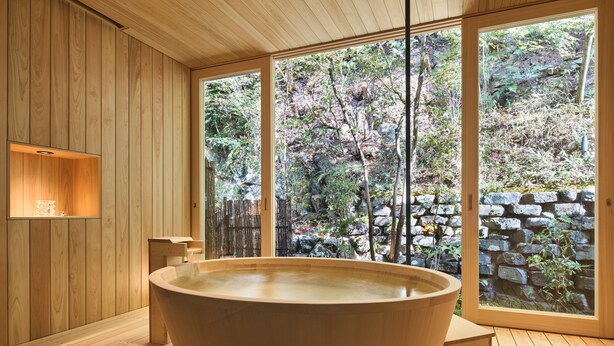 Interior de madera con una bañera grande, paredes de madera y vista a un jardín rocoso a través de amplias ventanas.