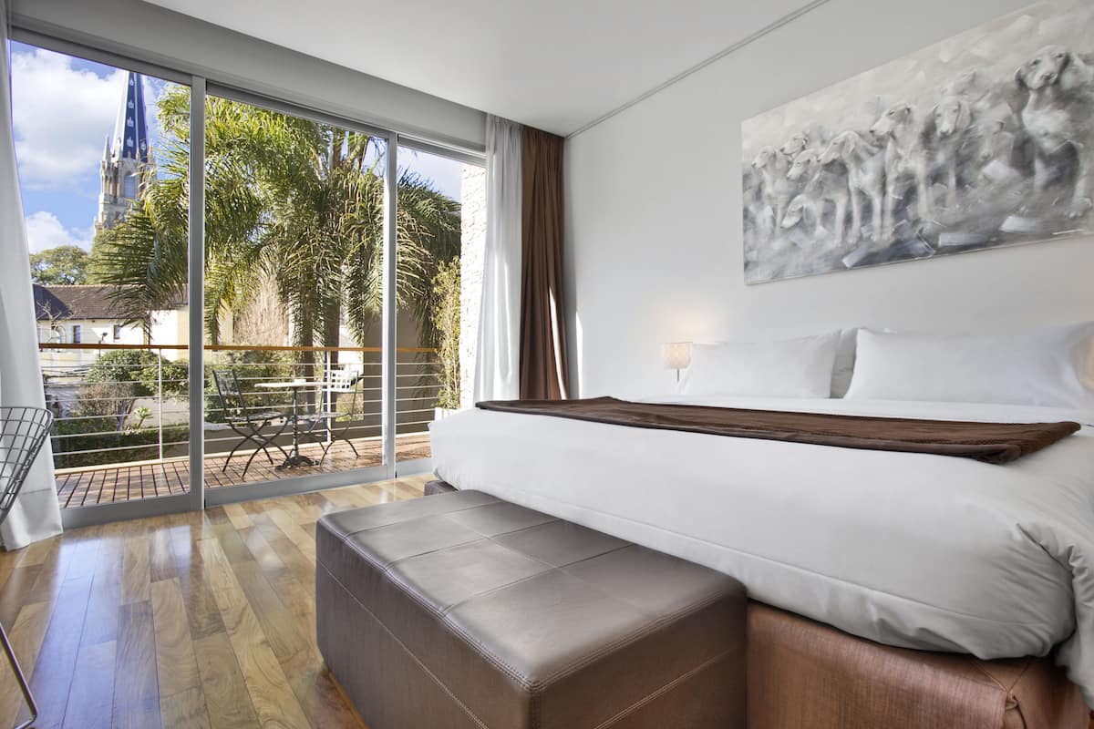 A hotel room with a large bed, a bench, a painting, and a view of a church and palm trees.