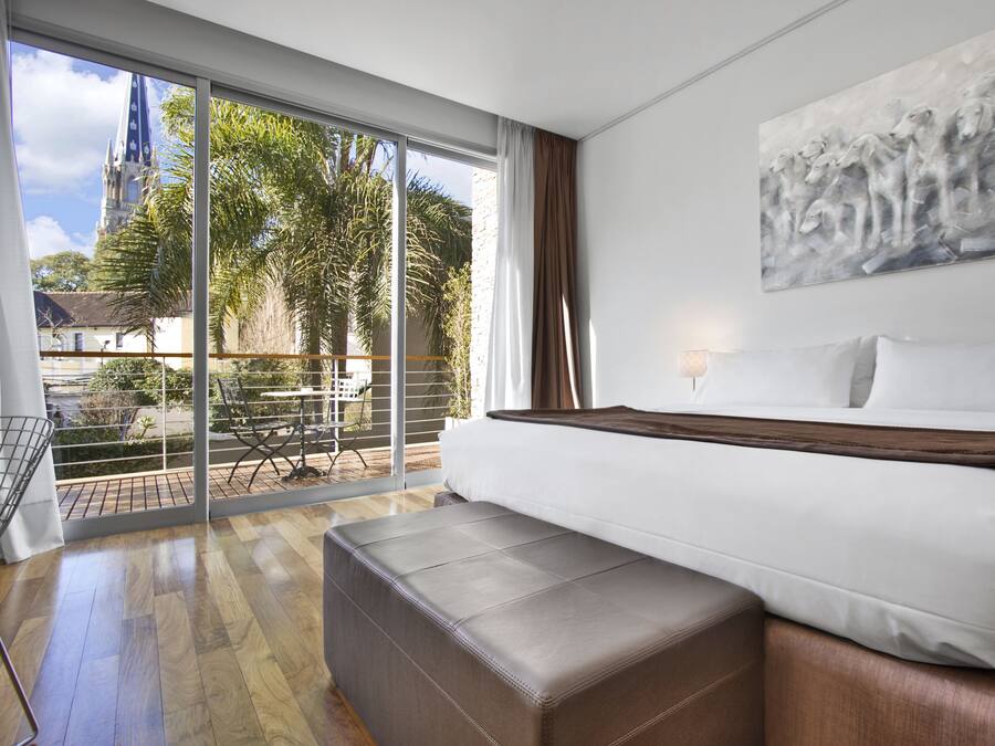 A hotel room with a large bed, a bench, a painting, and a view of a church and palm trees.