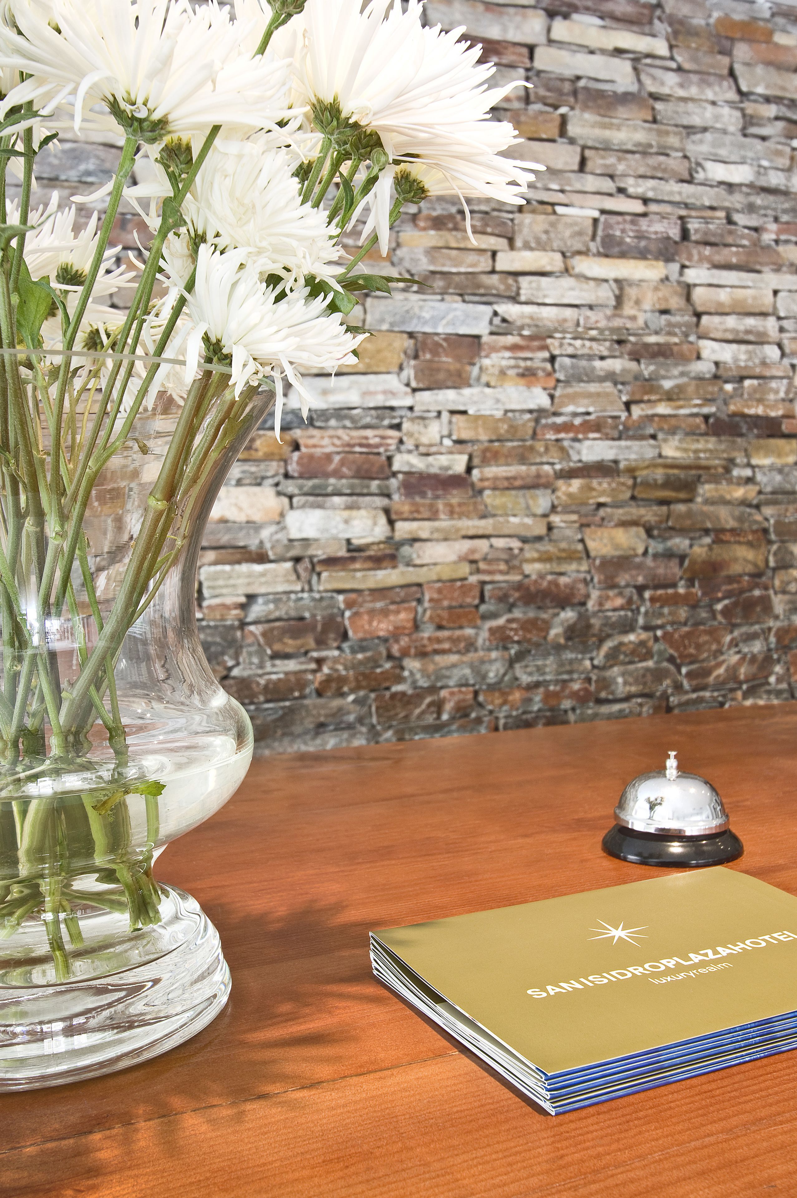 Un florero de cristal transparente con flores blancas sobre una mesa de madera, un folleto de hotel apilado y una campana al fondo.