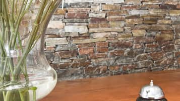 A clear glass vase with white flowers on a wooden table, a stack of a hotel brochure, and a bell in the background.