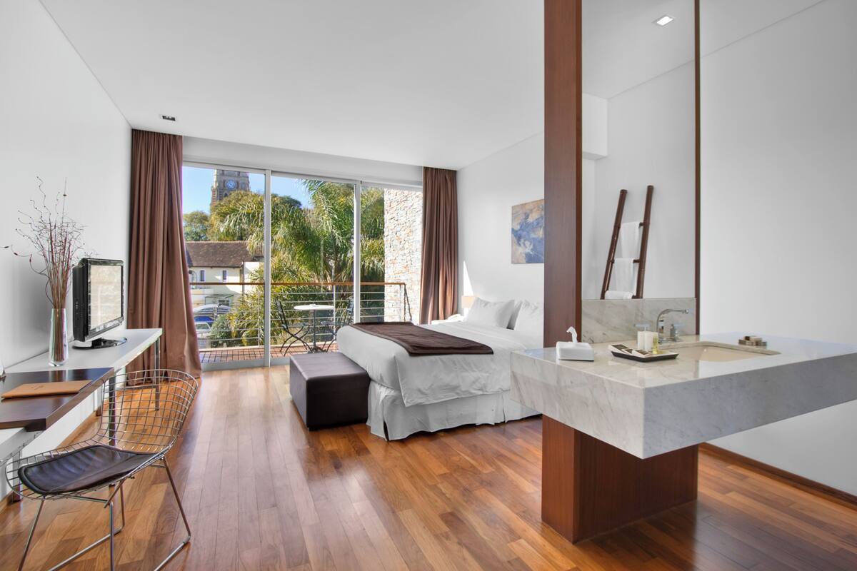 A modern hotel room with a bed, a desk, a TV, a balcony with a view of buildings and trees, and a bathroom with a large sink and mirror.