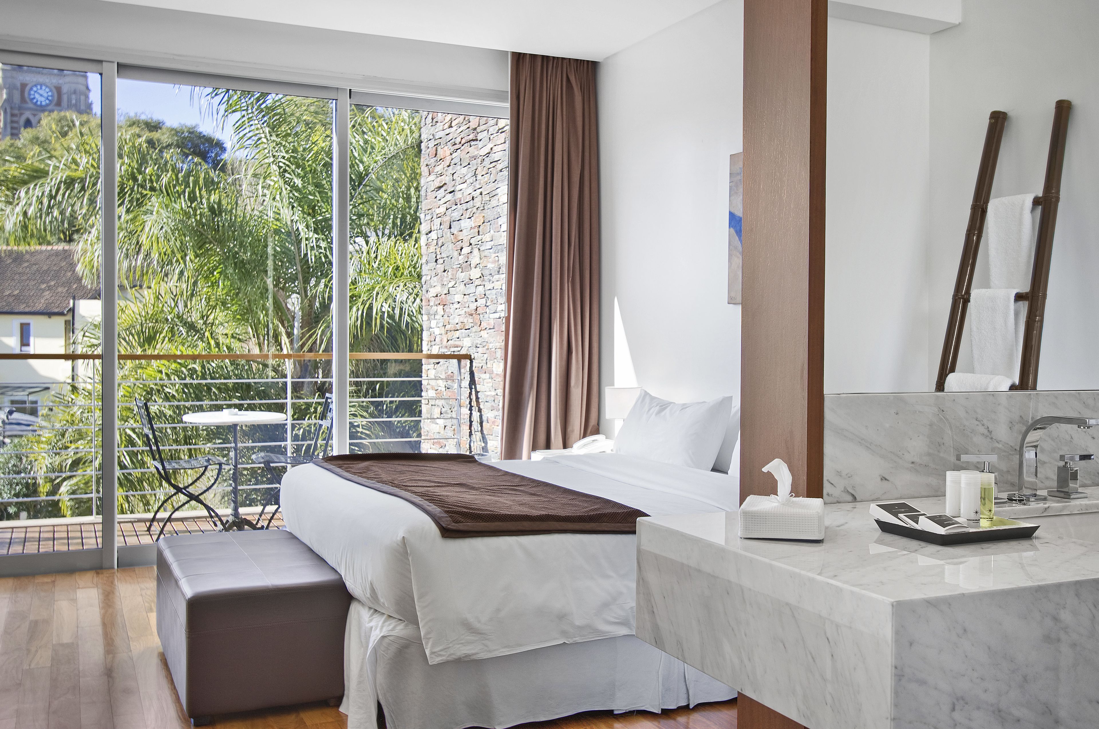 Quarto de hotel com varanda, uma cama, banheiro com bancada de mármore e vista para uma torre do relógio.