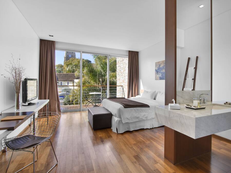 Quarto de hotel moderno com cama, escrivaninha, televisão, varanda com vista para edifícios e árvores, e banheiro com pia grande e espelho.
