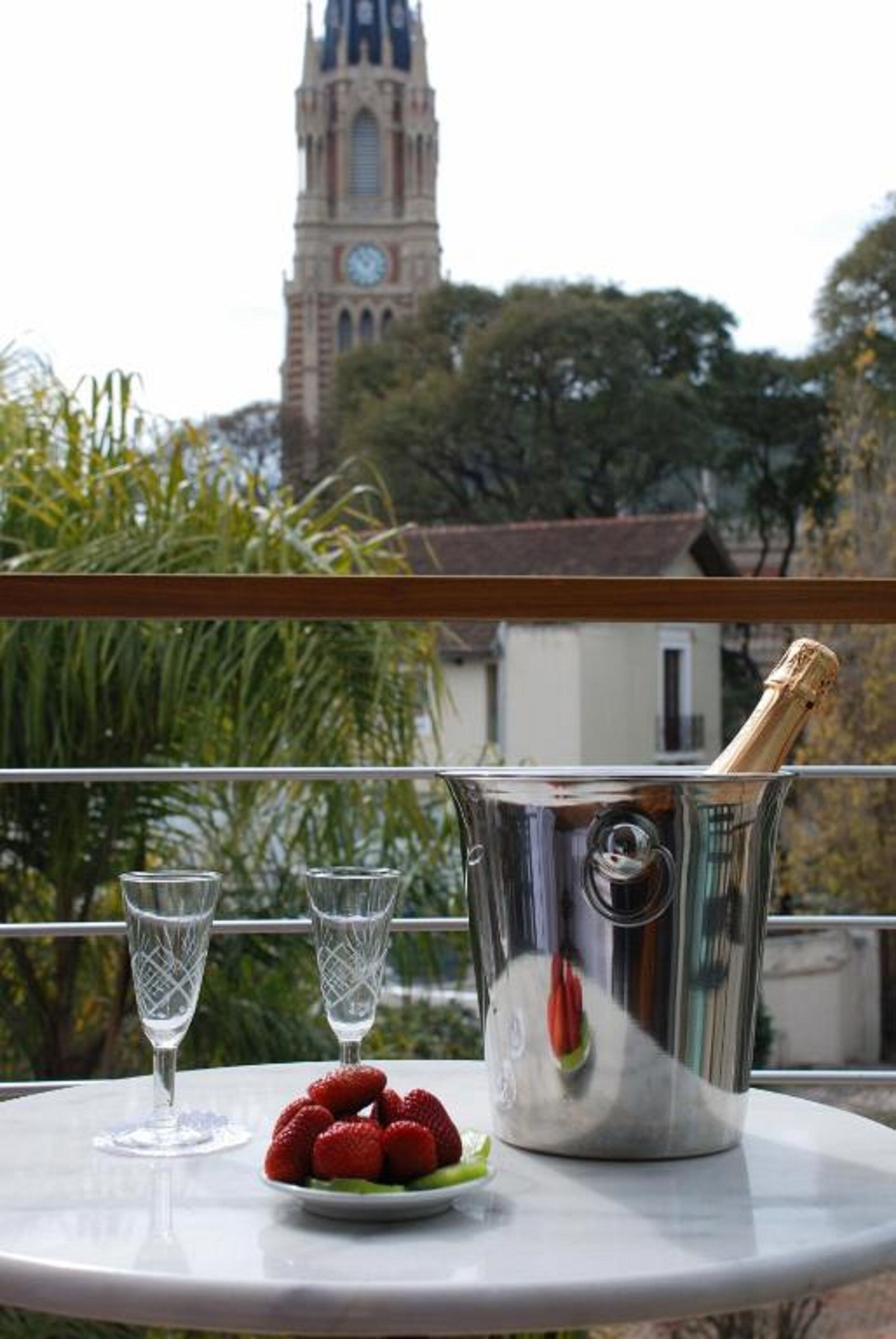 Un balcón con una mesa dispuesta para dos personas, que incluye una botella de champán, dos copas de vino y un plato de fresas. Se observa una torre de reloj histórica al fondo.