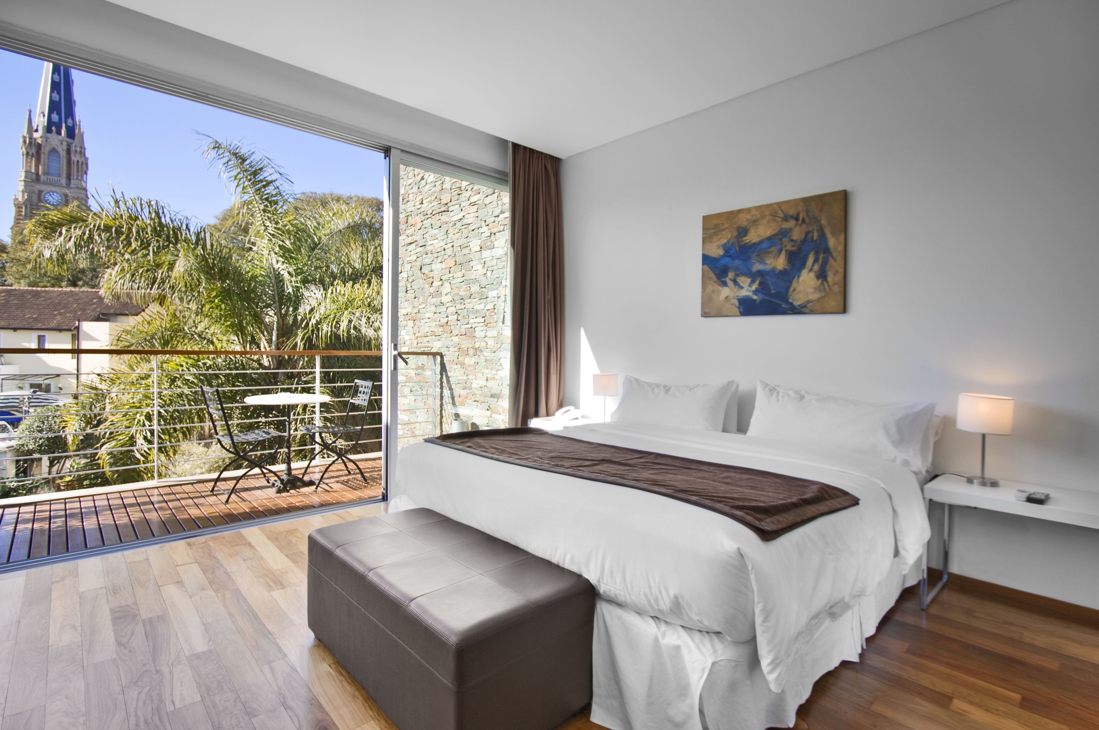 A bedroom with a large bed, a bench, a nightstand, a painting, and a view of a church and greenery.