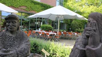 Steinstatuen in einem Gartenbereich mit Außenbestuhlung und einem Gebäude im Hintergrund.