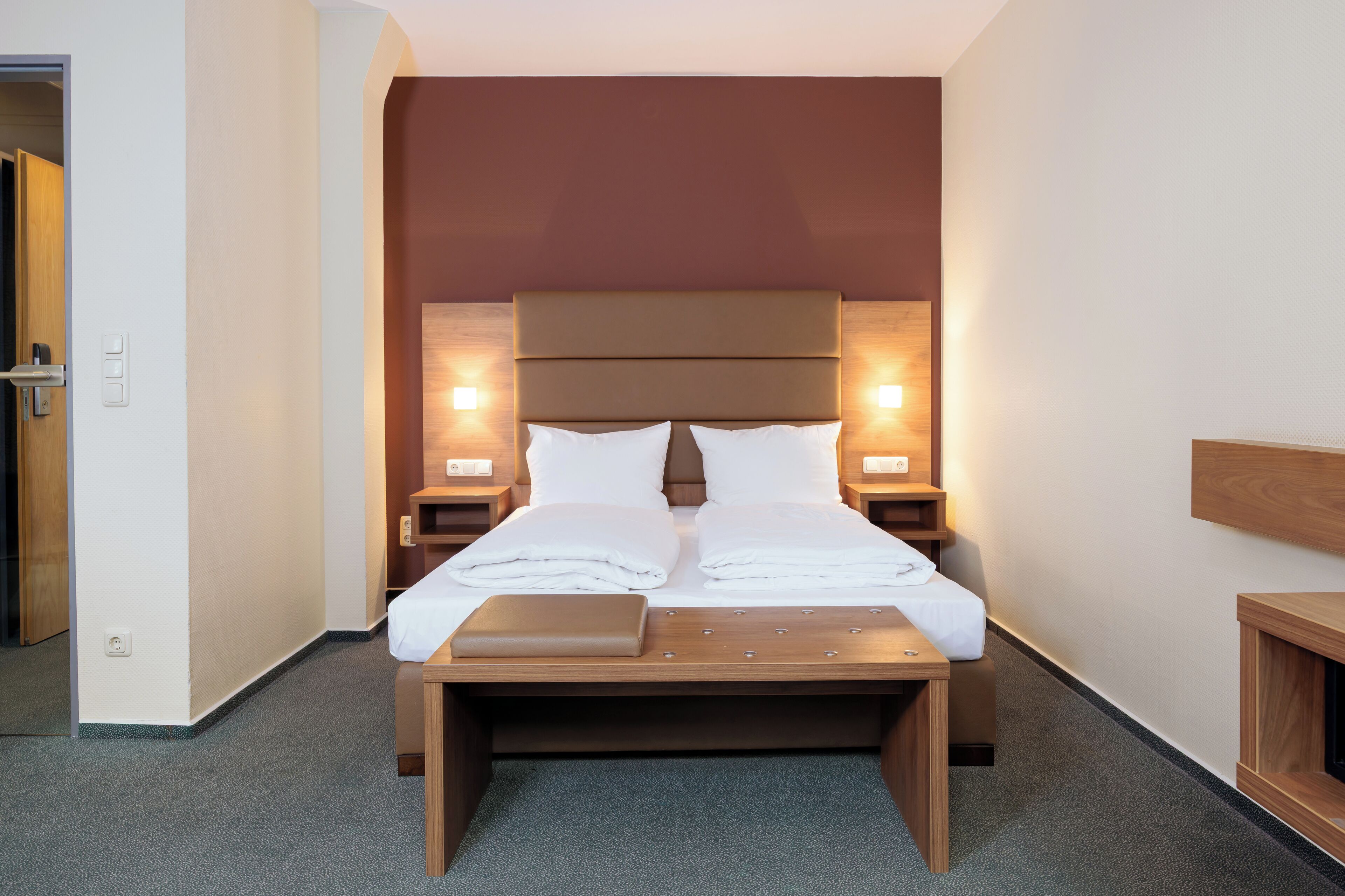 A neatly made bed with a wooden headboard, two bedside tables with lamps, and a small wooden bench at the foot of the bed.