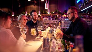 A bar with people seated at tables, holding drinks, and a bartender behind the bar. - GenoHotel Forsbach (Roesrath)